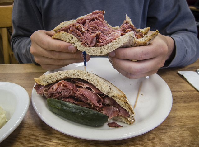 ein sehr grosses durchgeschnittenes Pastramisandwich mit einer halbe Pickled Gurke, zwie Hände halten einen Teil des sandwiches hoch, man kann sehr viel Pastrami erkennen