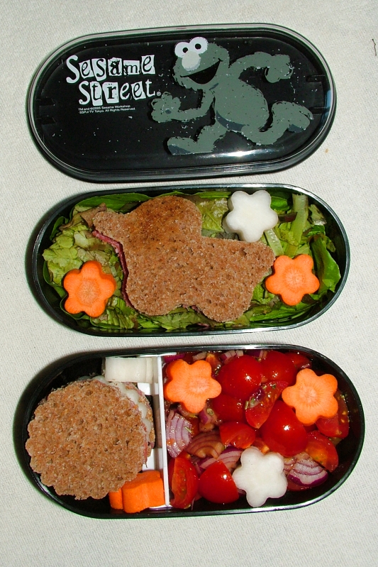 eine ovale schwarze Bentobox mit Deckel, oben der Deckel mit Elmo, in der Mitte ein als Ente ausgestochenes Brot auf Salatbett mit Möhre und Rettich als Blüte, unten ein Kirschtomatensalat mit roter Zwiebel, mehr Möhrenrettichblüten, und ein kleines rundes Sandwich